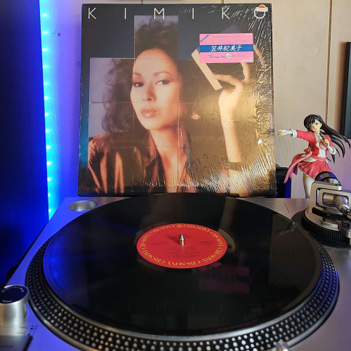 A vinyl record sits on a turntable. Behind the turntable, a vinyl album outer sleeve is displayed. The front cover shows a puzzle like theme around Kimiko Kasai doing a duck lips sort of look at the camera. She is holding up a block of wood with an image on it that is another piece of the puzzle