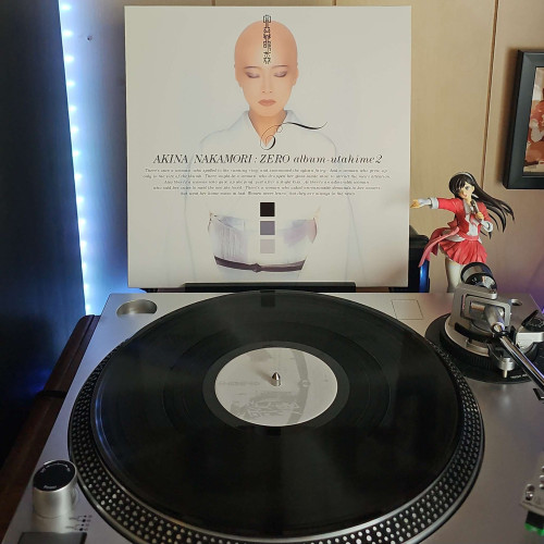 A vinyl record sits on a turntable. Behind the turntable, a vinyl album outer sleeve is displayed. The front cover shows a bald Akina Nakamori w/ her eyes closed wearing a kimono. 