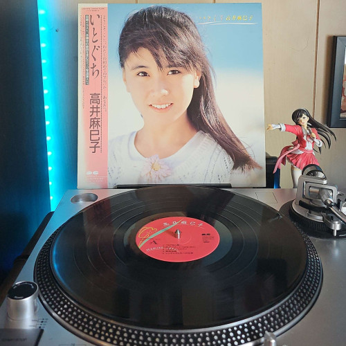 A vinyl record sits on a turntable. Behind the turntable, a vinyl album outer sleeve is displayed. The front cover shows Mamiko Takai from the shoulders up, looking at the camera. Behind her is a blurry background of a beach