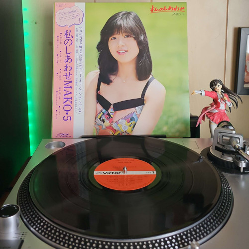 A vinyl record sits on a turntable. Behind the turntable, a vinyl album outer sleeve is displayed. The front cover shows Mako Ishino sitting in a field as she looks and smiles at the camera..