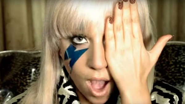 Lady Gaga. Her hair is blonde, long,with bangs and she has a lightning bolt painted on one cheek coming out of her right eye, Her  left eye is covered by the left hand. She is singing.