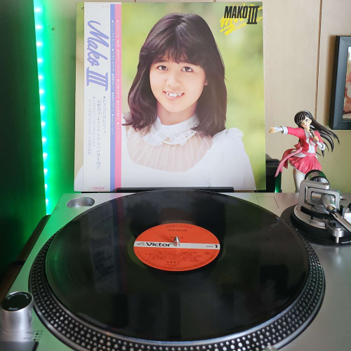 A vinyl record sits on a turntable. Behind the turntable, a vinyl album outer sleeve is displayed. The front cover shows Mako Ishino from the chest up, as she smiles at the camera. Behidn her is blurred out foliage.