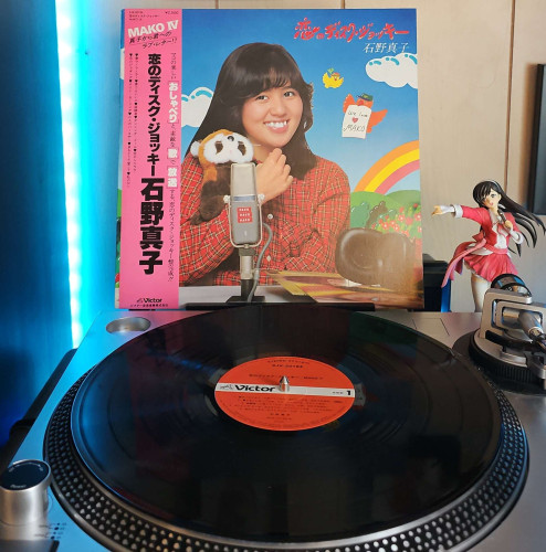 A vinyl record sits on a turntable. Behind the turntable, a vinyl album outer sleeve is displayed. The front cover shows Mako Ishino sitting at a desk smiling and hugging a red panda stuffed animal. On the desk are a microphone, headset, and vinyl records. 