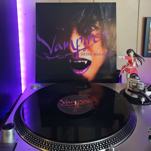 A vinyl record sits on a turntable. Behind the turntable, a vinyl album outer sleeve is displayed. The front cover shows Akina Nakamori's face. She has her mouth open and vampire fangs. 