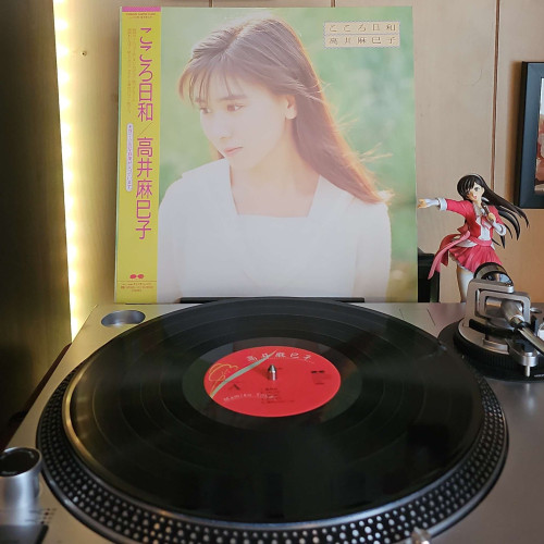 A vinyl record sits on a turntable. Behind the turntable, a vinyl album outer sleeve is displayed. The front cover shows Mamiko Takai looking down to the left. The background is a blurred field behind her. 