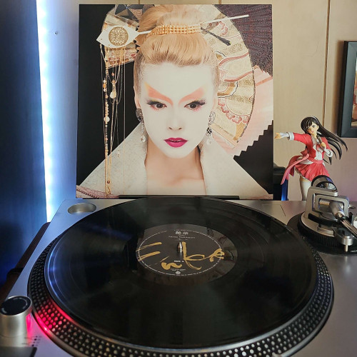 A vinyl record sits on a turntable. Behind the turntable, a vinyl album outer sleeve is displayed. The front cover shows a headshot of Akina Nakamori done up in geisha like makeup, wearing traditional style head pieces