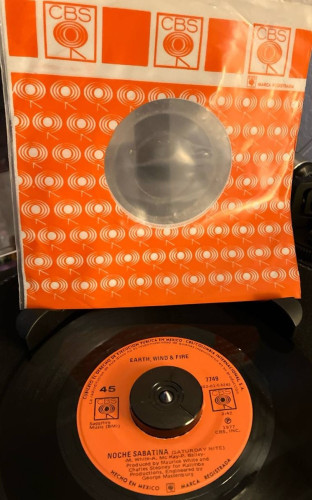 Seven inch record on a turntable with an orange label with black text. "Earth Wind & Fire* at the top. CBS logos on both sides of the spindle hole. "Noche Sabatina (Saturday Nite)" and credits on the bottom. Above the record a plastic orange sleeve with a repeated CBS pattern.