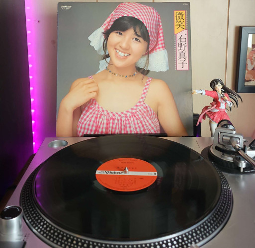 A vinyl record sits on a turntable. Behind the turntable, a vinyl album outer sleeve is displayed. The front cover shows Mako Ishino in a pink plaid top and headwear. 
