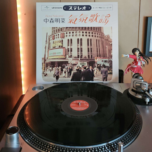 A vinyl record sits on a turntable. Behind the turntable, a vinyl album outer sleeve is displayed. The front cover shows a vintage image from Japan of a large building and people walking around. 