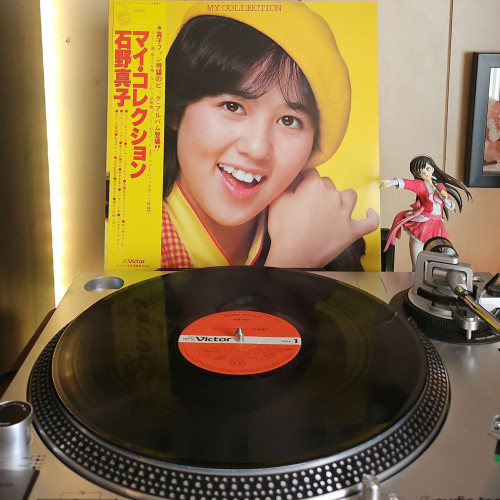 A vinyl record sits on a turntable. Behind the turntable, a vinyl album outer sleeve is displayed. The front cover shows Mako Ishino from the shoulders up. She is wearing a cap and smiling at the camera. 